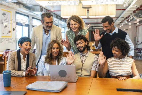 Diverse business colleagues at office desk having an online video conference, waving hands and smiling at laptop screen, collaborating and communicating remotely