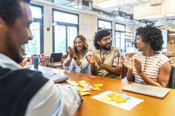 Diverse business colleagues sitting at a table in a modern office, happily clapping and smiling during a successful project meeting or new achievement