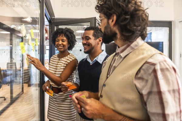 Diverse business team collaborating in a modern office, sharing ideas and planning strategy by arranging colorful sticky notes on a glass wall during a creative brainstorming session