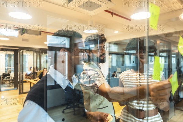 Business colleagues collaborating on a project, writing ideas on sticky notes affixed to a glass wall in a modern office, fostering teamwork and strategic planning in a dynamic coworking environment