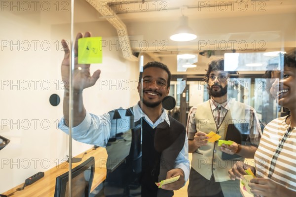 Happy diverse business team collaborating on glass wall during brainstorming session, sharing ideas and strategizing using colorful sticky notes in a contemporary workspace
