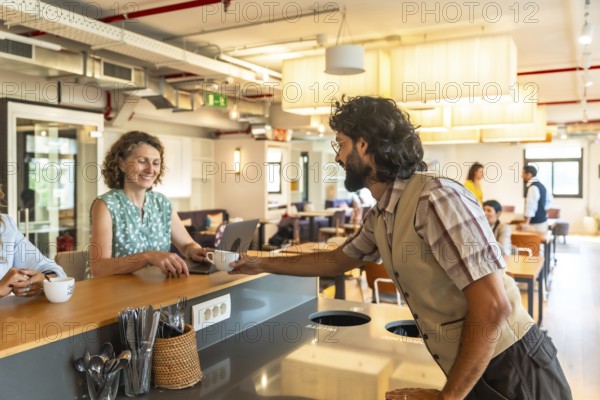 Young man passing a cup of coffee to a smiling woman working on her laptop at a counter in a modern, brightly lit coworking space, with other colleagues socialising in the background
