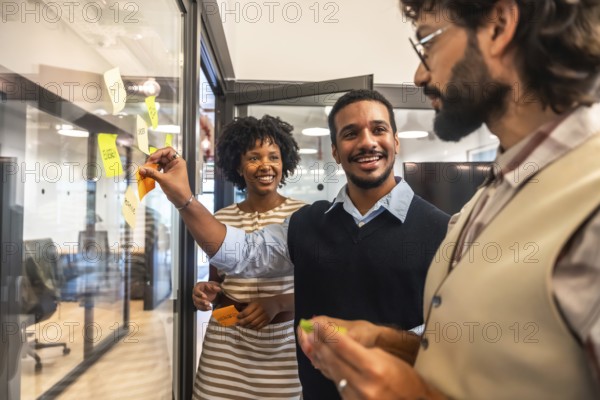 Diverse business colleagues collaborating on a project, smiling and applying colorful sticky notes to a glass wall during a creative brainstorming session in a modern office