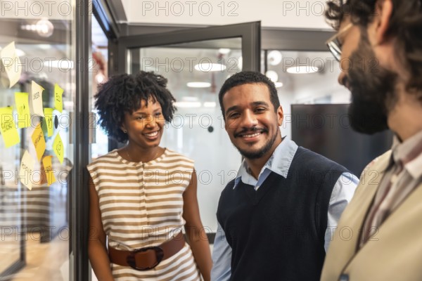 Coworkers smiling and engaging in discussion, brainstorming ideas on a glass board with sticky notes, representing teamwork, diversity, and successful business collaboration