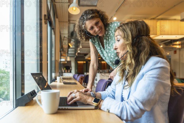 Two women wearing headsets are working together at a desk by a window, with one colleague providing assistance and guidance to the other using a laptop in a coworking space