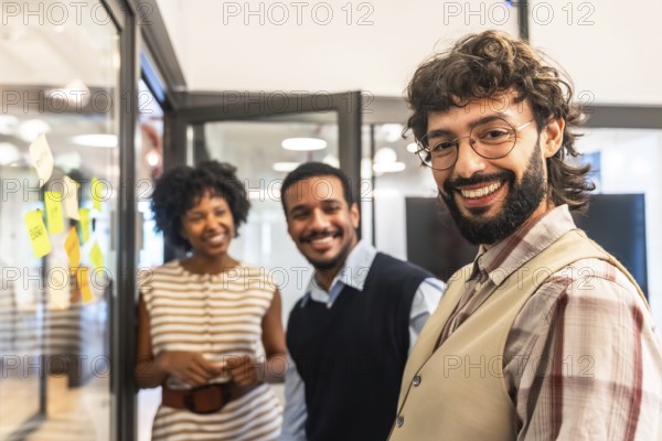 Group of diverse colleagues standing together and smiling in a contemporary coworking office, showcasing teamwork and a positive business environment with sticky notes on a glass wall
