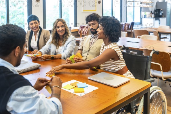 Diverse business team, including a woman in a wheelchair, collaborating around a wooden table in a modern coworking space, brainstorming and sharing ideas energetically
