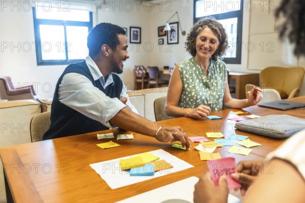Diverse coworkers sitting around a wooden table, smiling and collaborating on new project ideas using colorful sticky notes during a brainstorming session in a modern office