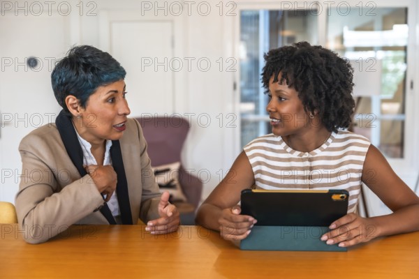 Two diverse women colleagues collaborating and talking about business while sitting at a table in an office or coworking space, one holding a digital tablet