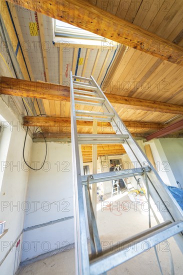 View of an attic with a ladder leading to an open window. Wooden ceiling elements and sunlight dominate the room