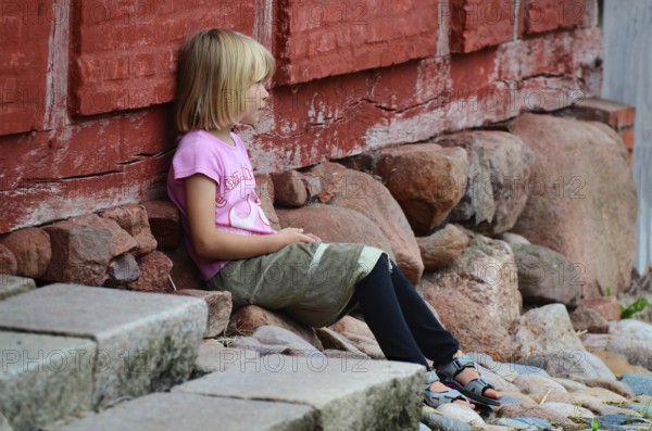 Little blonde girl sitting next to a red half-timbered house and looking to the side in Den gamle by, The old Funen village, open air museum in Odense, Fyn island, Funen, Denmark, Scandinavia