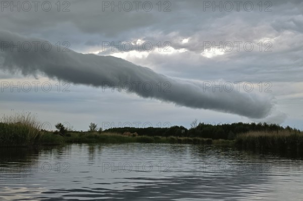 A long cloud tube stretches across a calm lake with reeds and cloudy sky, Peenetal nature park Park, Mecklenburg-Western Pomerania, Germany