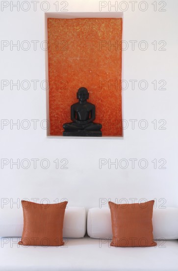 Statue of a black Buddha sitting meditating against an orange background, including bed with pillows and castors, Palace Hotel Devigarh, near Udaipur, Rajasthan, India