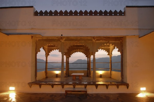 View through illuminated, traditional arched architecture of a slightly elevated terrace of mountains lying in the warm light of dusk, Palace Hotel Devigarh, Delwara village near Udaipur, Rajasthan, India