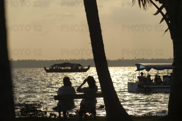 A couple sitting on a lakeside bench at sunset, boats gliding in the water, romantic atmosphere, Coconut Lagoon Hotel, Cgh Earth Group, Lake Vembanad, Kerala, India