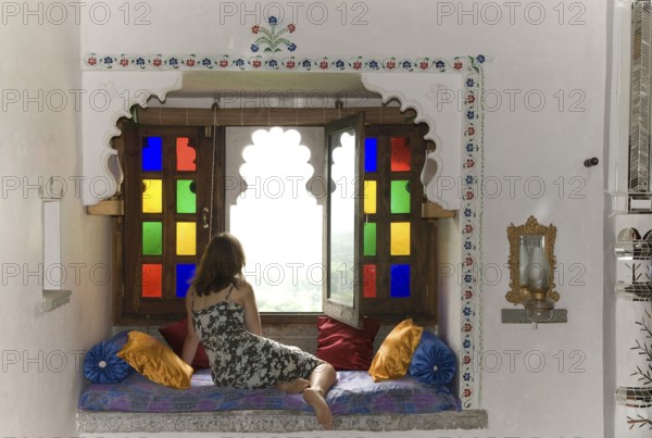 Woman wearing a dress in a cozy sitting area with pillows in front of colorful window, Palasthotel Karni Fort Bambora, Bambora, Rajasthan, India