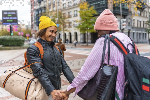 Multiethnic gay couple holding hands, beanies and backpacks, smiling as they walk through a vibrant city street with luggage, enjoying an autumn urban travel moment