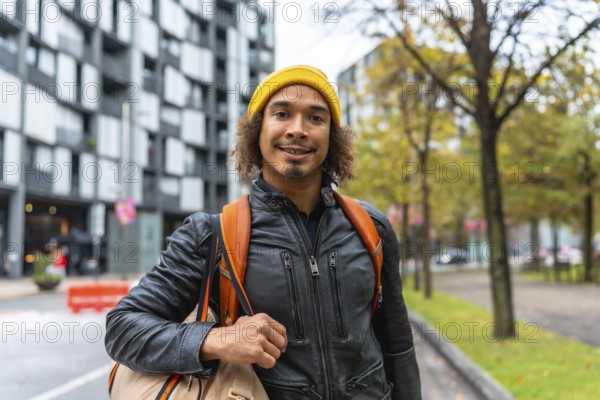 Young man with curly hair in a yellow beanie and leather jacket, backpack on his shoulders, smiling confidently while walking along a modern city street on a crisp day