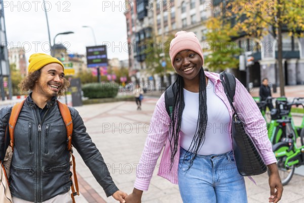 Young multiracial couple smiling and holding hands while strolling down a city street lined with autumn trees, enjoying a casual, cozy outdoor date and relaxed urban togetherness