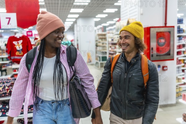 Diverse young couple smiling while holding hands, walking together down an aisle in a retail store during holiday shopping for christmas presents and decorations