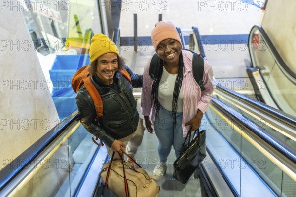Happy young couple moving up an escalator at an airport or train station, carrying their travel bags and backpacks, looking up and smiling confidently at the camera