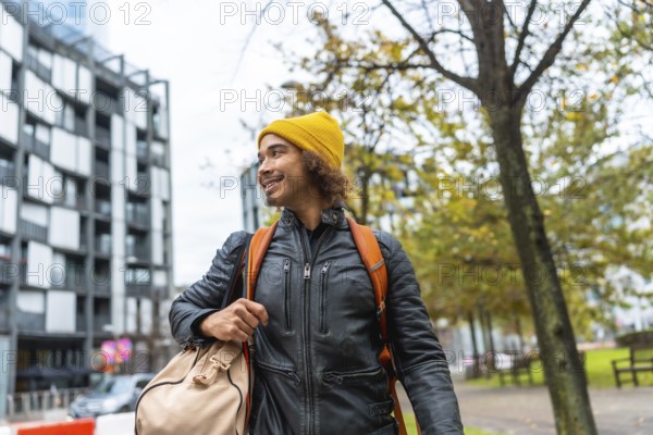 Young man with a yellow beanie and black leather jacket carrying a duffel bag and backpack, smiling while walking through an urban park with autumn trees and modern buildings