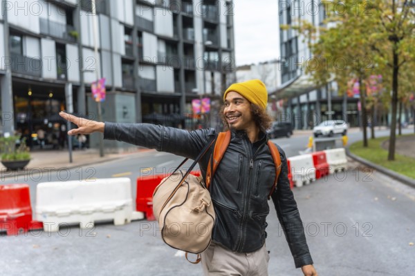 Happy young multiracial man wearing a yellow beanie and leather jacket pointing with his hand while walking and carrying a backpack on an urban street with modern buildings