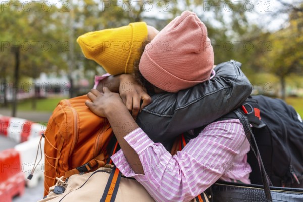 Two young adults are sharing a close, emotional embrace outdoors, holding onto each other with suitcases and bags suggesting travel, departure, or reunion