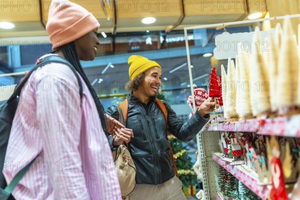 Young Mixed-race couple happily shopping for festive holiday decorations, selecting small christmas trees and ornaments on store shelves, celebrating the seasonal spirit together