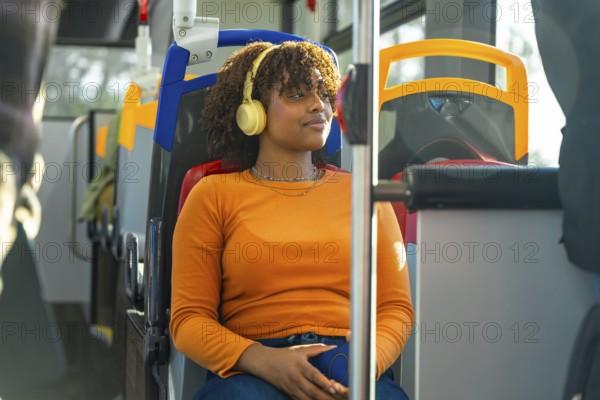 Young black woman with afro headphones relaxes and listens to music on a city bus during her commute, gazing out the window, peaceful and immersed in her journey