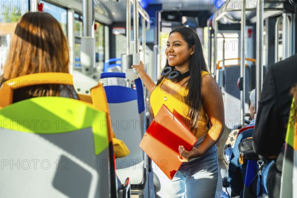 Young woman smiling, standing inside a modern public transportation bus, holding an orange binder and headphones around her neck, commuting to or from studies or work