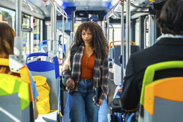 Young woman passenger with long curly hair and glasses walking along the aisle of a public bus, carrying a laptop and a backpack, surrounded by vibrant seat colors