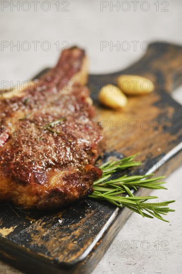 A mouthwatering grilled steak is placed on a weathered wooden cutting board. Fresh rosemary and caramelized garlic cloves are arranged beside it, creating an appealing and savory presentation