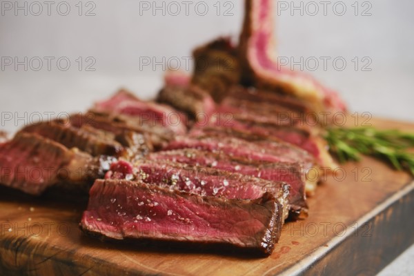 Close-up view of sliced grilled t-bone steak medium rare on a wooden cutting board. The meat is perfectly cooked with a juicy pink center. Fresh herbs and seasoning add color and flavor to the presentation
