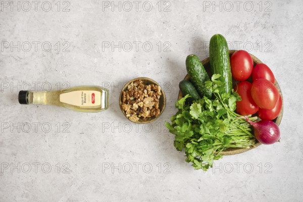 A colorful arrangement of fresh tomatoes, cucumbers, and herbs on a light gray surface. A bowl of crunchy walnuts and a bottle of wine vinegar complete the group, ideal for salad making