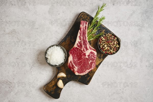 A fresh cowboy steak on a wooden cutting board, surrounded by garlic cloves, coarse salt, and a bowl of mixed peppercorns. Rosemary sprigs add a touch of color, ideal for grilling