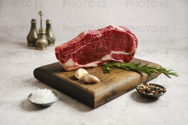 Raw ribeye roll steak on a wooden cutting board surrounded by garlic cloves, fresh rosemary, salt, and mixed peppercorns. The scene highlights the ingredients for a delicious meal