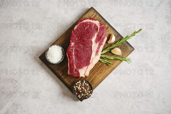 Raw ribeye roll steak rests on a wooden cutting board alongside garlic cloves and fresh rosemary. Nearby, bowls contain sea salt and a mix of colorful peppercorns, ideal for seasoning