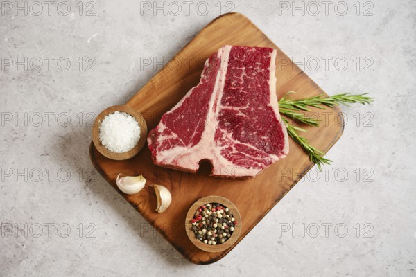 A large, marbled raw t-bone steak on a wooden cutting board surrounded by garlic cloves, salt, mixed peppercorns, and fresh rosemary. The scene highlights a culinary preparation setting