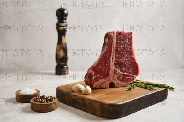 A thick cut of raw t-bone steak on a wooden cutting board with cloves of garlic, fresh rosemary, and bowls of salt and pepper. This setup invites preparation for a delicious meal