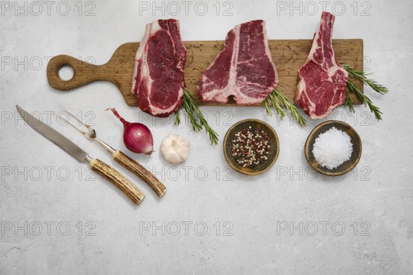 Three thick cuts of beef steaks on a wooden chopping board with fresh rosemary, salt, pepper, onion and garlic