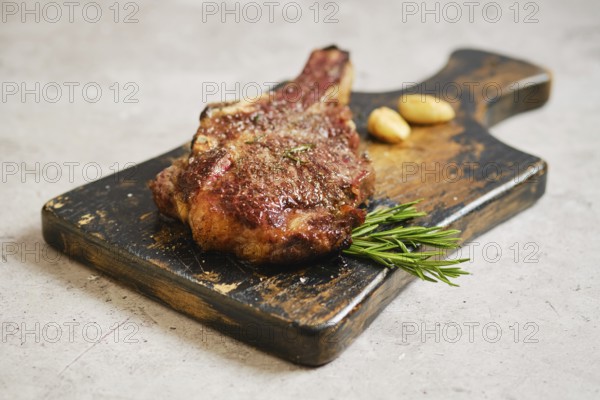 A perfectly cooked cowboy steak on a rustic wooden cutting board, garnished with fresh rosemary and roasted garlic cloves. This appetizing scene showcases a delicious meal ready to be enjoyed