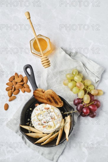 A cheese platter on a light background, showcasing a brie cheese wheel drizzled with honey. Almonds, a slice of bread, and fresh grapes surround the cheese