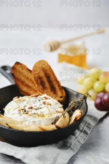 A baked cheese dish is elegantly presented in a cast iron skillet alongside toasted bread and fresh fruits. Honey in a jar adds a sweet touch to this appetizing setup, perfect for a cozy gathering