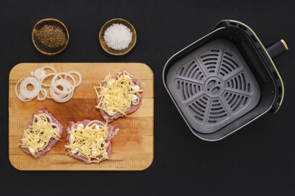 Raw pork chops are topped with grated cheese and sliced onions on a wooden cutting board. Nearby, bowls contain spices like salt and pepper, along with an air fryer basket awaiting the meal prep