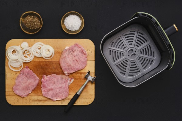 Raw pork chops on a wooden cutting board next to mallet. Sliced onions are ready beside them, with an air fryer basket waiting for the meal preparation to begin