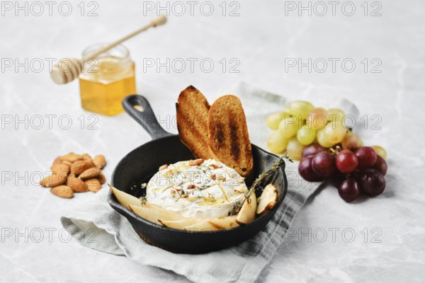 A cast iron skillet holds warm baked cheese, garnished with herbs. Nearby are toasted bread slices, almonds, grapes, and a honey jar, creating an inviting spread for sharing and enjoyment