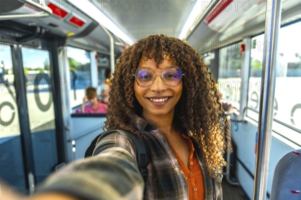 Young woman with curly hair and glasses smiling at camera while taking a happy selfie on a city bus, enjoying public transit during a daytime urban commute
