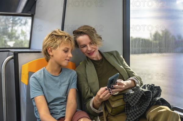 Mother and son sharing a smile as they browse a smartphone together on a city bus, enjoying a casual moment of connection and modern family bonding during their commute