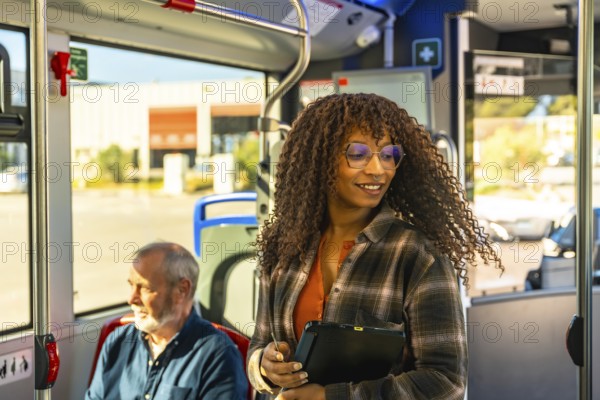 Woman with glasses smiles while standing on a city bus, holding a digital tablet among commuters during a bright daily commute, modern, eco friendly urban travel